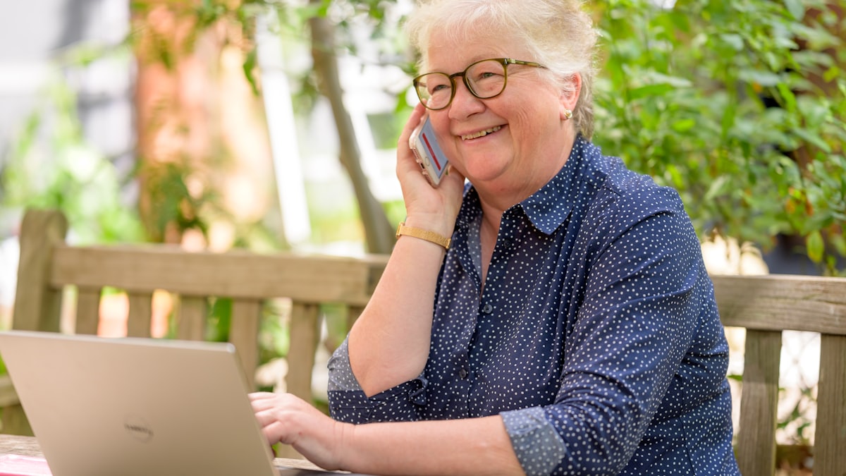 Older woman in glasses smiling while talking on a smartphone outdoors with a laptop